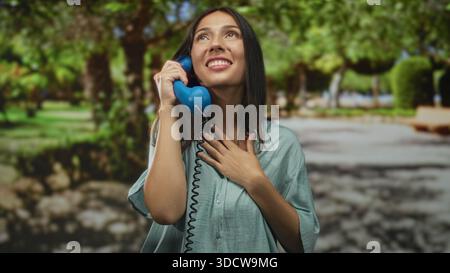 Woman holds blue telephone receiver to ear while hand rests over chest in park; gratitude connection. Stock Photo