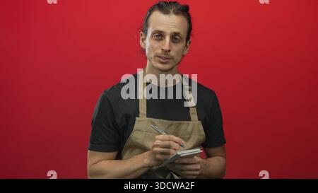 Man with long hair tied back wearing an apron and black tshirt writing on a small notepad with a pen while looking down in a red studio; concentration Stock Photo