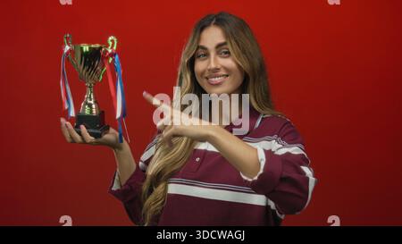 Woman holds trophy in studio with finger pointing to trophy highlighting the cup against red background; achievement celebration success pride. Stock Photo