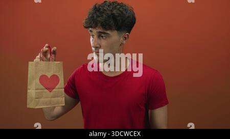 Young man wearing a red shirt holding a brown paper bag with a red heart and shushing while standing in orange studio; secrecy. Stock Photo