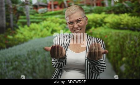 Woman beckons with both hands and visible palms on a forest path near green foliage, smiling at camera in striped shirt and glasses; welcoming invitat Stock Photo