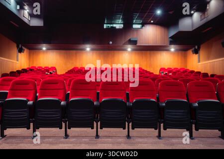 Empty modern auditorium with rows of red upholstered seats, warm wooden acoustic wall panels, ceiling spotlights and balcony, ready for cinema screeni Stock Photo