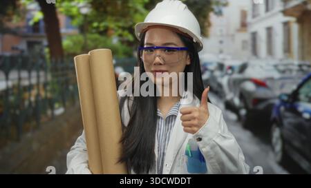 Chinese woman architect wearing hardhat holds blueprints and gives thumbs up on a vibrant street, symbolizing professionalism and confidence in an urb Stock Photo