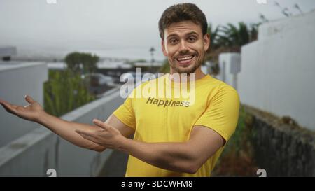 Man posing with hands on hips, wearing yellow tshirt printed happiness, smirking slightly beside a white wall and cactus on a street; happiness. Stock Photo