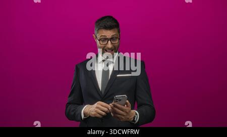 Man in business suit and tie tapping a smartphone with both hands while reading the screen in magenta studio; concentration professionalism. Stock Photo