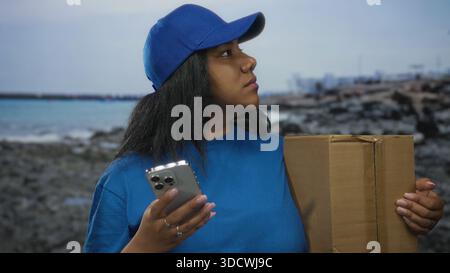 Delivery courier woman in blue uniform holds smartphone and a cardboard box while standing on a rocky beach seaside shore; dedication. Stock Photo