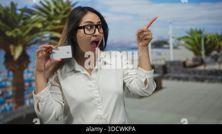 Woman holding white creditcard near face and points finger toward beach promenade with palm trees while wearing glasses and open mouth; surprise savin Stock Photo