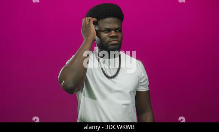 Man touching his ear with hand while wearing white t shirt and wooden bead necklace against magenta wall in studio; doubt. Stock Photo