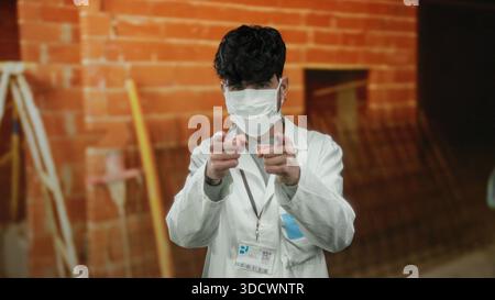 Young man in mask and lab coat at construction site points at camera, indicating attention or focus, blending professional attire with industrial sett Stock Photo
