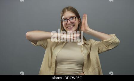 Young blonde woman wearing glasses smiles while framing face with hands in grey studio; playful joy. Stock Photo