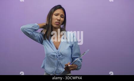 Thai woman in blue blouse holding clipboard with hand on neck showing neck pain in studio; work stress concern. Stock Photo