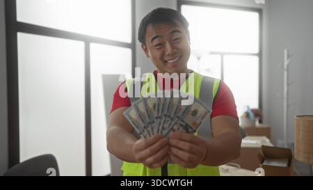 Young man holding money in new home interior wearing safety vest, showcasing happy expression amidst moving boxes, embodying success and construction Stock Photo