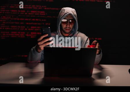 Dangerous hooded fraudster in glasses sitting at desk staring at smartphone while holding credit card, laptop open and green code reflecting on wall Stock Photo