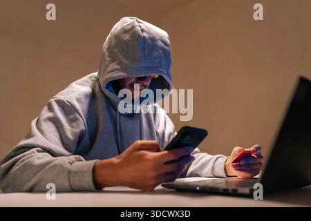 Hooded person engaging in cybercrime, performing online fraud and data theft using mobile phone, laptop and credit card in dark environment Stock Photo