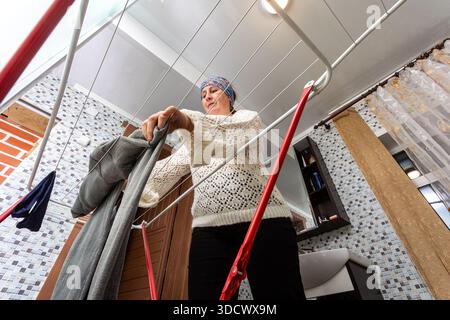 Hanging washed laundry on wire clothes dryer in utility room of country house, middle aged woman does household chores. Stock Photo