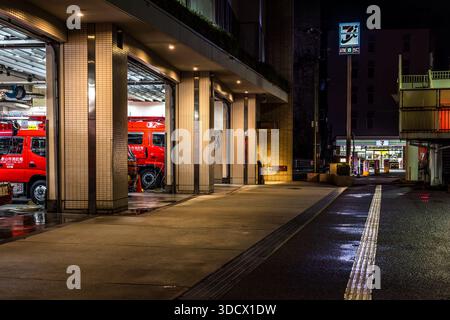 A red fire engine of the Wakayama fire department stands in the open vehicle bay of a fire station in Wakayama, Japan, with the illuminated sign of a 7-Eleven store visible in the background. Rain-wet asphalt reflects the street lighting. Stock Photo