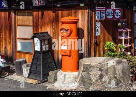An old orange cylindrical postbox stands in front of a wooden building in the old town of Yuasa, Wakayama Prefecture, Japan, beside a wooden lantern box and a boulder used as a seat. Yuasa is regarded as the birthplace of Japanese soy sauce, and the historic quarter has been a protected preservation district since 2006. Stock Photo