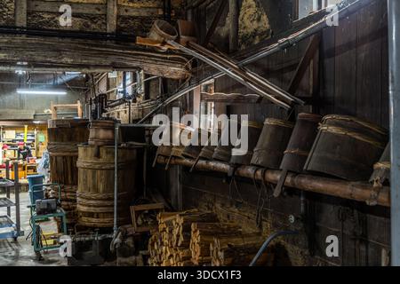 Wooden barrels and stacked firewood stand in a traditional soy sauce factory in Yuasa, Wakayama Prefecture, Japan, metal pipes and a wooden funnel hanging above the equipment. Yuasa is regarded as the birthplace of Japanese soy sauce, produced here for centuries with similar tools. Stock Photo