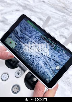 Man’s hands holding a drone remote controller with a screen showing an aerial view of a snowy landscape. Modern technology concept and travel. Stock Photo