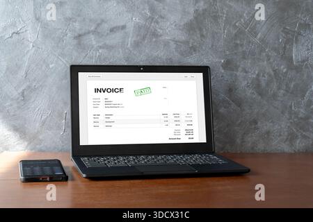 Laptop showing a paid invoice on a wooden desk with a smartphone calculator. Business finance concept for billing, accounting, payment confirmation, o Stock Photo