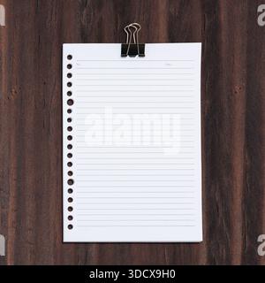 Top view of a white, lined paper sheet with hole punches on the left side, attached to a dark wood background with a black binder clip. Perfect for mo Stock Photo