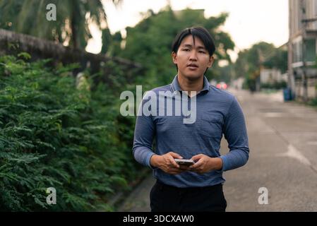 Young asian man standing outdoors holding smartphone, browsing internet Stock Photo