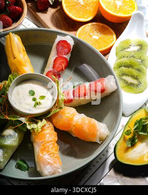 Delicious and healthy fruit spring rolls served with a creamy white dip, placed on a stylish ceramic plate surrounded by sliced fruits Stock Photo
