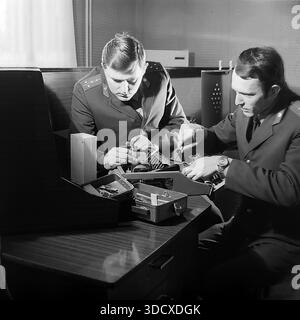 Ukraine, Donetsk region, Sloviansk, 1980s. An archival black and white photograph capturing two Soviet militia officers working at a desk inside the Department of Private Security. The uniformed men are focused on repairing or adjusting a complex electronic device, likely an alarm system or radio equipment, using hand tools. This authentic image documents the technical side of law enforcement work and daily service routine in a peaceful Donbas before the war, preserving history. Stock Photo