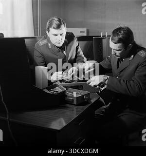 Ukraine, Donetsk region, Sloviansk, 1980s. An archival black and white photograph capturing two Soviet militia officers working at a desk inside the Department of Private Security. The uniformed men are focused on repairing or adjusting a complex electronic device, likely an alarm system or radio equipment, using hand tools. This authentic image documents the technical side of law enforcement work and daily service routine in a peaceful Donbas before the war, preserving history. Stock Photo