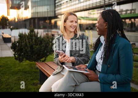 Diverse businesswomen relaxing on bench laughing enjoying conversation Stock Photo