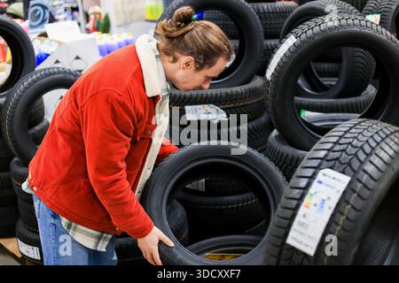 Young man with bun hairstyle in red jacket choosing car tires in auto store, inspecting rubber wheel for purchase. High quality photo Stock Photo