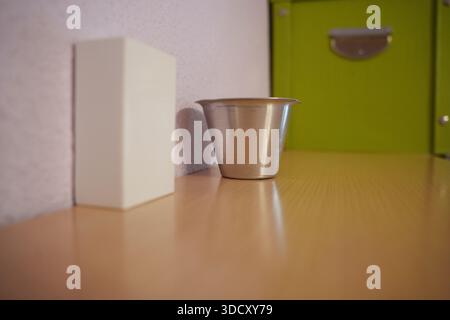 Small metal cup on wooden countertop with white box and green drawer in soft focus, film grain texture, warm ambient light, minimalist domestic scene, Stock Photo