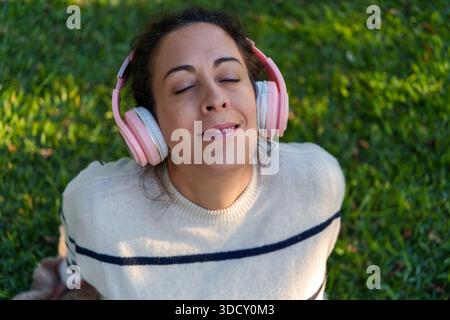 Woman relaxing on green grass, smiling with her eyes closed while listening to music through pink headphones, enjoying a peaceful moment Stock Photo