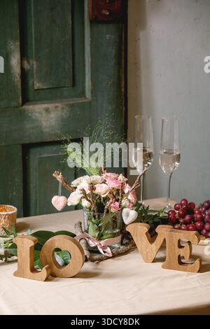 st. Valentines day or wedding romantic table setting with wooden letters love, needle felted hearts, bouquet of pink roses, couple glasses of champagne and red grapes on natural linen tablecloth. Stock Photo