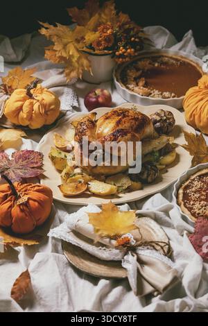 Thanksgiving or Halloween dinner with baked chicken with potatoes and lemons on big ceramic dish, pumpkin pies, plates, yellow autumn leaves and pumpkins as decorations over white table cloth. Stock Photo