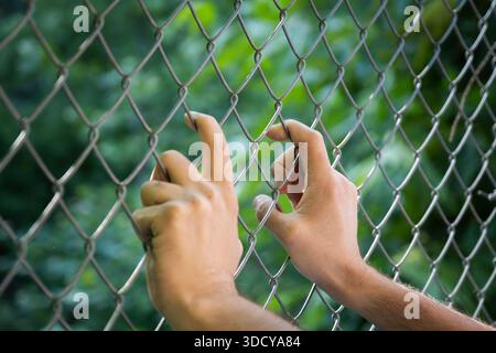 Human hands gripping a wire fence, symbolizing the refugee crisis, migration, and the desperate search for freedom, hope, and new opportunities beyond Stock Photo