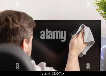 Rear view of a man's hand carefully wiping a large black desktop monitor with a soft cloth, demonstrating diligent cleaning and care for electronic de Stock Photo