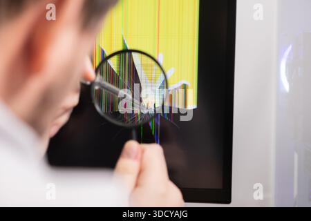 A service technician performs a meticulous inspection of a cracked computer monitor screen featuring vibrant pixel lines using a magnifying glass for Stock Photo