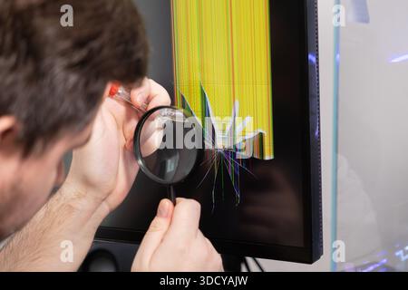 Close-up of a professional technician using a magnifying glass and a screwdriver to inspect a broken computer monitor display with colorful vertical l Stock Photo