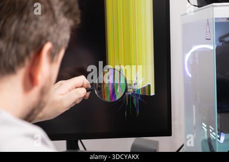 Close-up technician using magnifying glass to inspect cracked monitor showing colorful lines, emphasizing electronic troubleshooting and repair Stock Photo
