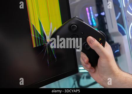 Close-up of an angry person's hand with a modern black game controller breaking a desktop computer screen, displaying colorful lines and illustrating Stock Photo