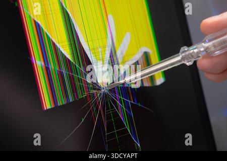Close-up of a human hand holding a transparent screwdriver, touching a heavily damaged electronic display with fractured glass and colorful pixel glit Stock Photo