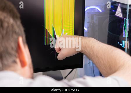 Angry person's fist strikes computer monitor, creating spiderweb cracks and bright vertical lines, conveying technology stress, frustration, and urgen Stock Photo