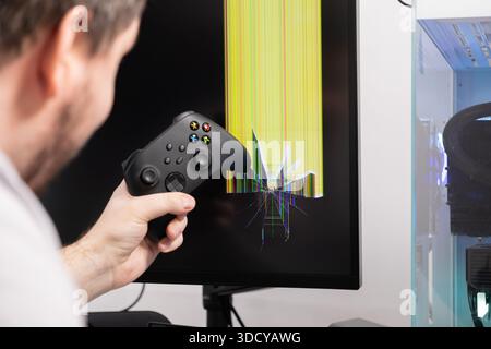 Gamer's hand holds console controller near gaming PC setup with critically damaged monitor displaying yellow lines and glass cracks, symbolizing hardw Stock Photo