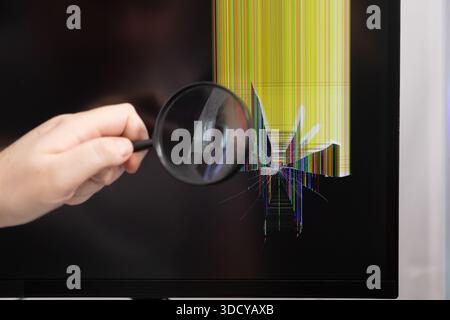 A persons hand using a magnifying glass to examine a shattered computer screen displaying vertical multicolored lines, illustrating device damage and Stock Photo
