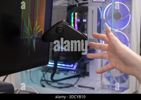 A black game controller floats mid-air as a hand reaches towards it, contrasting with a shattered monitor and a gaming PC, conveying gaming frustratio Stock Photo