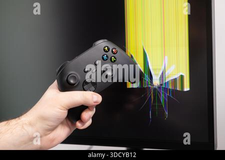 Close-up of a person's hand holding a modern black video game controller, positioned against a severely cracked monitor displaying colorful digital da Stock Photo
