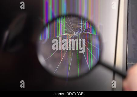 Close-up of a magnifying glass held over a broken LCD or LED display featuring starburst cracks and multicolored vertical pixels, highlighting device Stock Photo