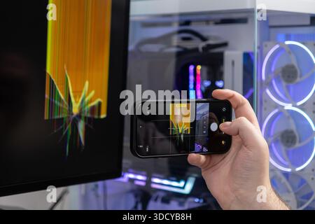 Close-up of a hand holding a smartphone capturing the damage on a broken computer monitor with colorful vertical lines, illustrating necessary IT diag Stock Photo