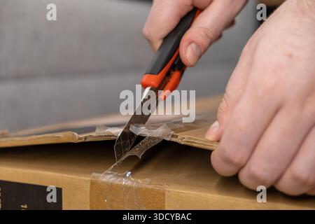 Close-up view of human hands using a utility knife to slice open a taped brown cardboard parcel, illustrating the process of receiving an e-commerce o Stock Photo